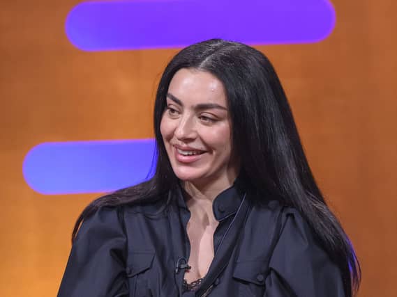 Charli XCX in a leather jacket smiling while sitting on a red couch